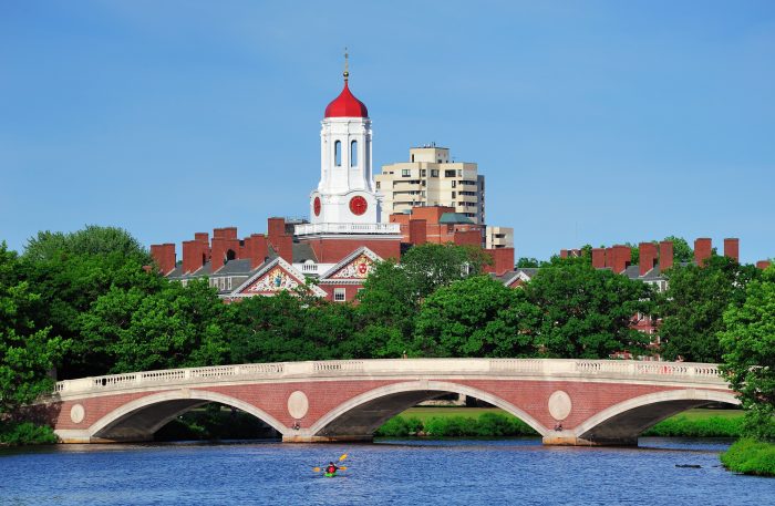 Harvard campus in Boston John W. Weeks Bridge and clock tower over Charles River in Harvard University campus in Boston with trees, boat and blue sky.